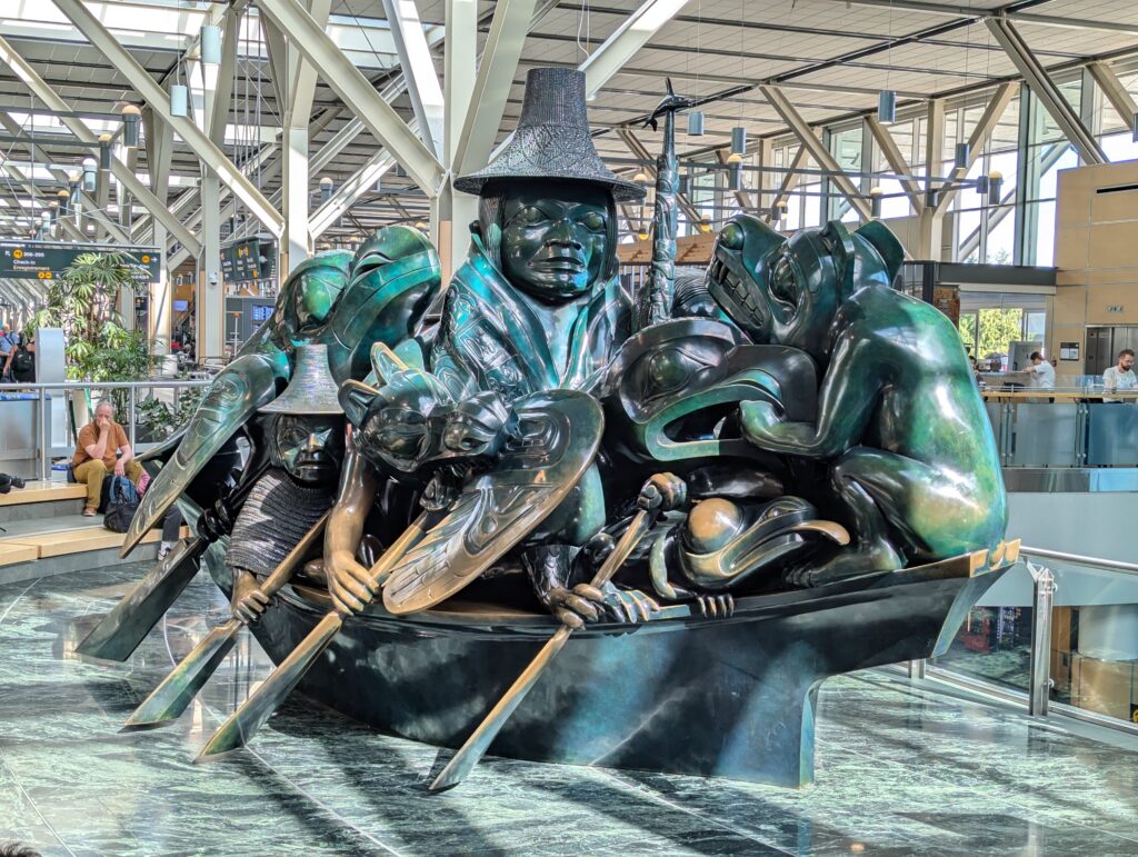 A photo of a large jade-like sculpture in the Vancouver International Airport. It depicts a Haida person in a dugout canoe surrounded by a variety of animals. The piece is titled "The Spirit of Haida Gwaii" by Bill Reid.