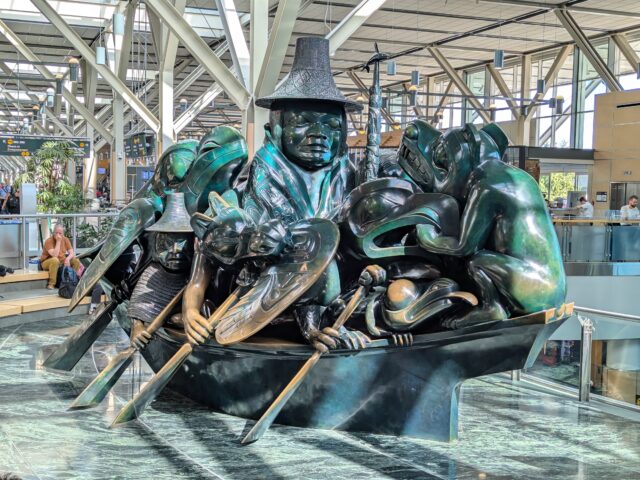 A photo of a large jade-like sculpture in the Vancouver International Airport. It depicts a Haida person in a dugout canoe surrounded by a variety of animals. The piece is titled "The Spirit of Haida Gwaii" by Bill Reid.